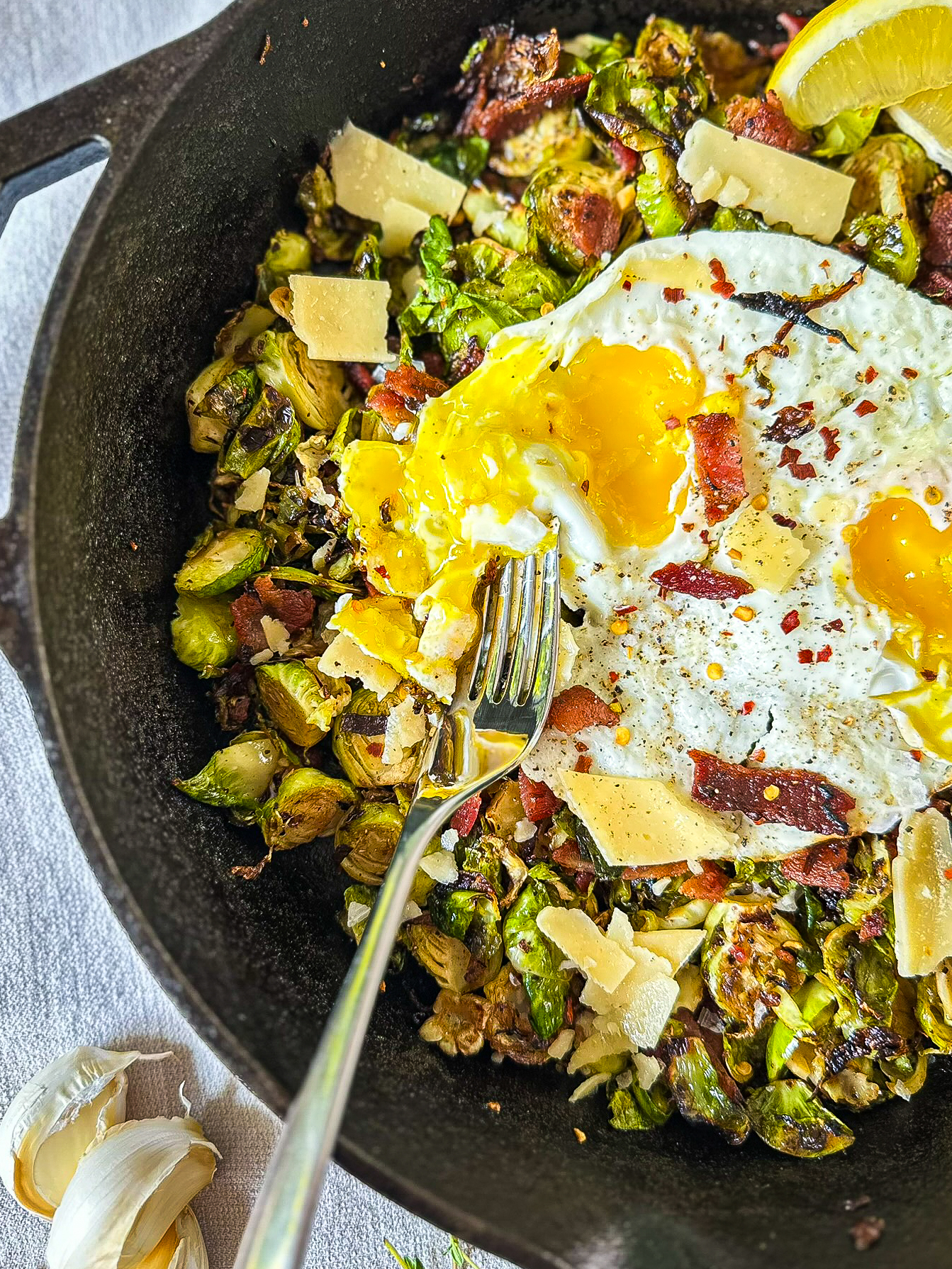 black skillet with a hash mixture of egg, brussels sprouts, bacon, shaved parmesan, lemon wedges, and a silver fork sitting on linen with garlic cloves