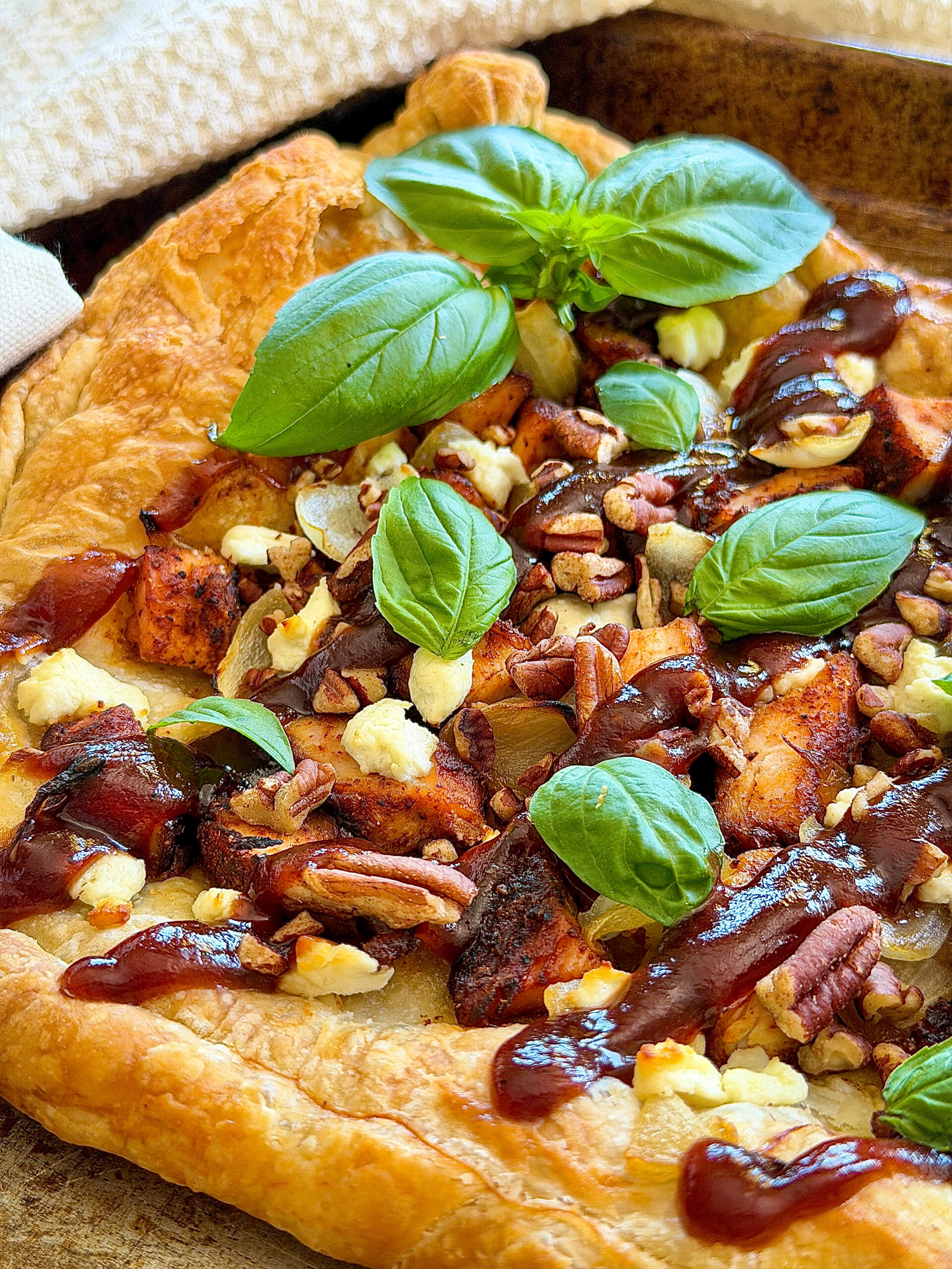 Pizza with BBQ sauce, diced chicken, goat cheese crumbles, onions, and fresh basil leaves sitting on brown pan with white towel