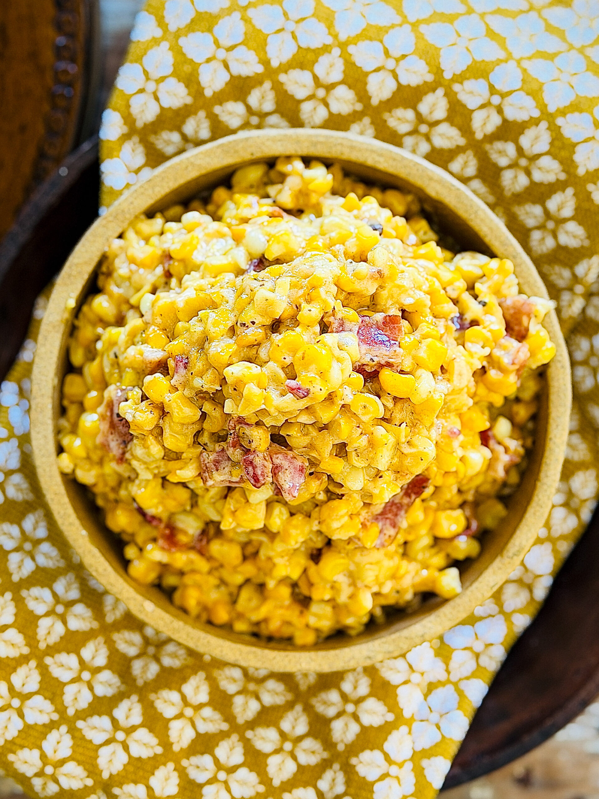 yellowigh tan bowl with heaping pile of creamed corn and bacon sitting on a mustard yellow and white patterned towel on a wood table