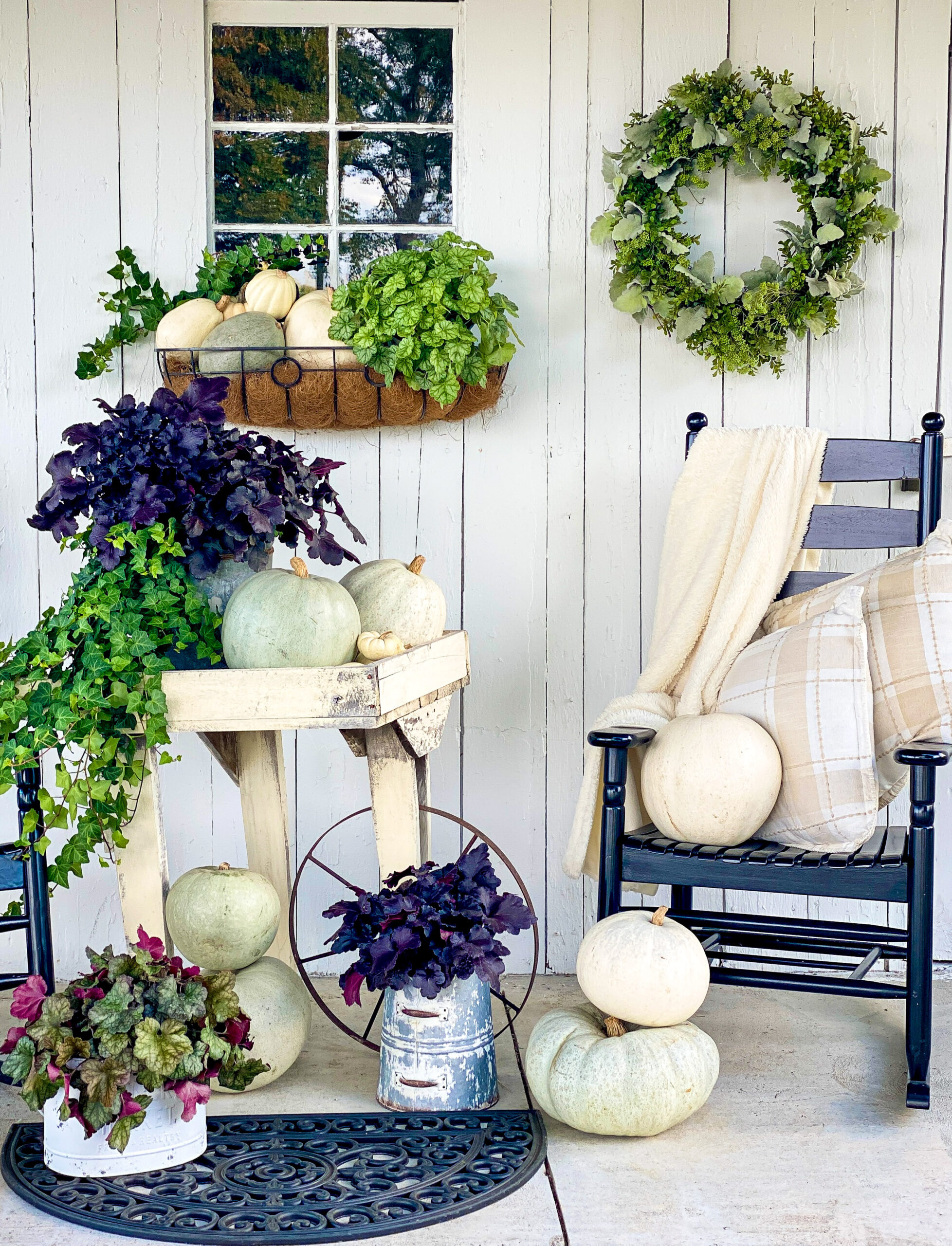 leafy green and deep purple plants with light green and white pumpkins sitting all around porch in from of white barn with window, black rocking chair with cream blanket and pillows and green wreath