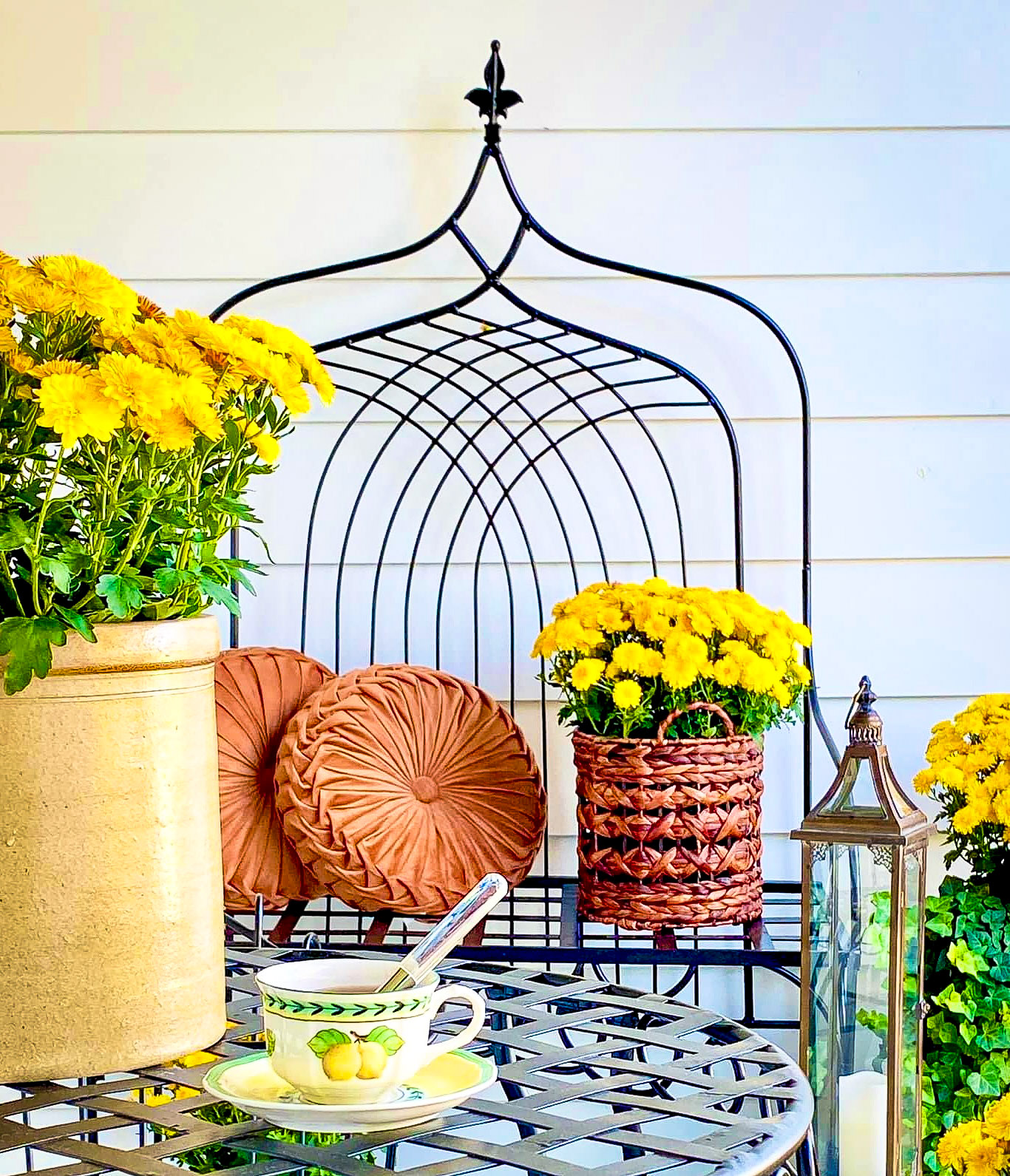 yellow flowers with green stems in ceramic and wicker pots with decorative iron table and bench, brown round pillows, tall glass lamp, and tea cup with lemons