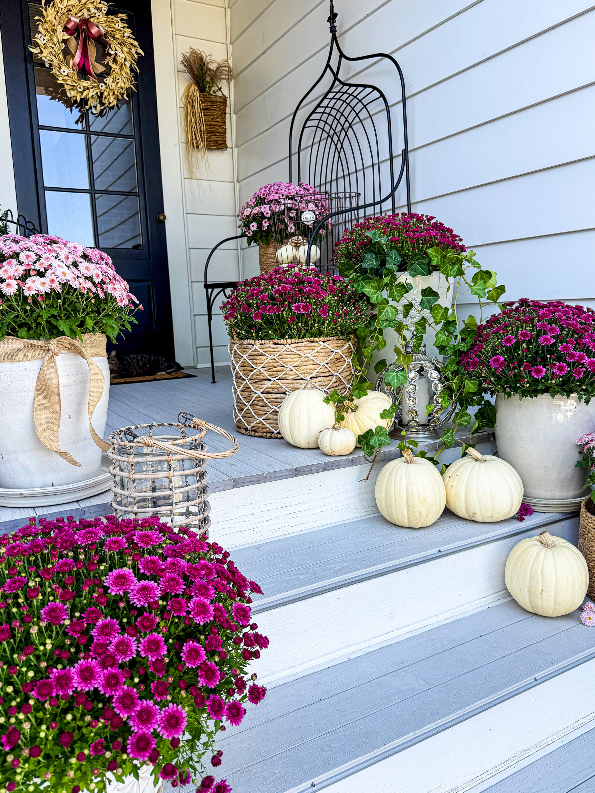 maroon and light pink flowers with green leaves sitting on light gray and white porch and steps, with white pumpkins and black door in background with brown wreath