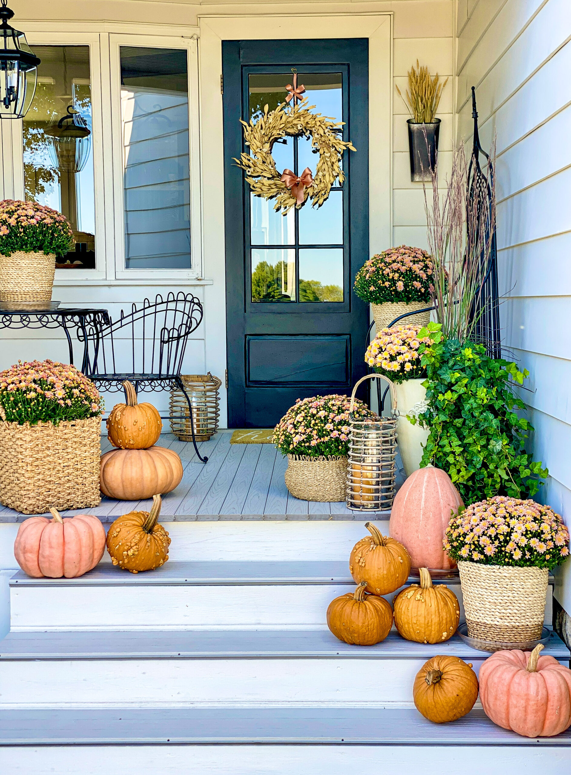 orange pumpkins and pinkish orange flowers with green stems sitting in wicker pots on wood porch with black door and tan wreath