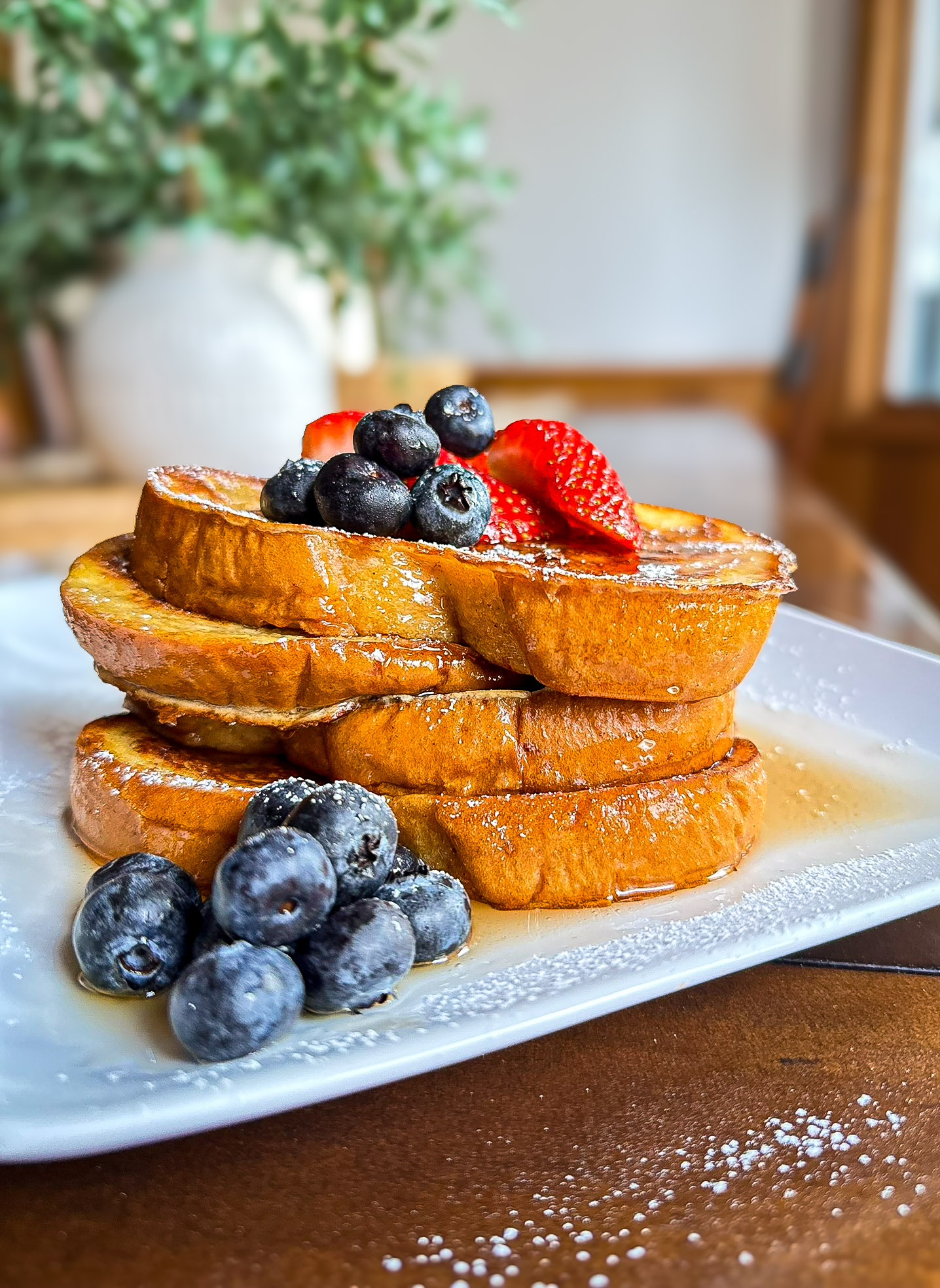 white plate of frech toast slices with syrup, powdered sugar, blueberries, and strawberries with green plant in white vase in background
