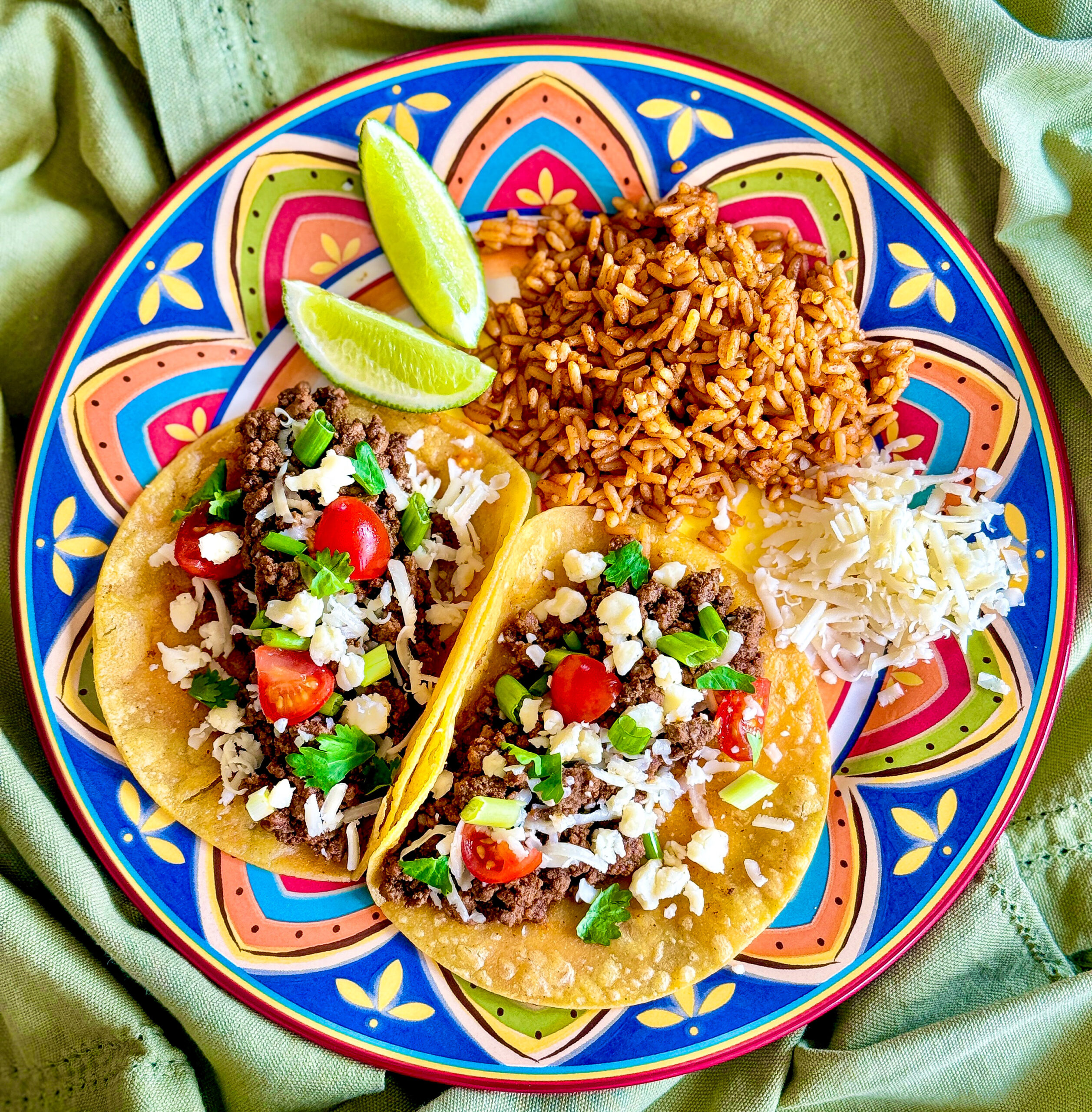 close up of festive blue design plate with tacos, rice, lime wedges sitting on light green cloth
