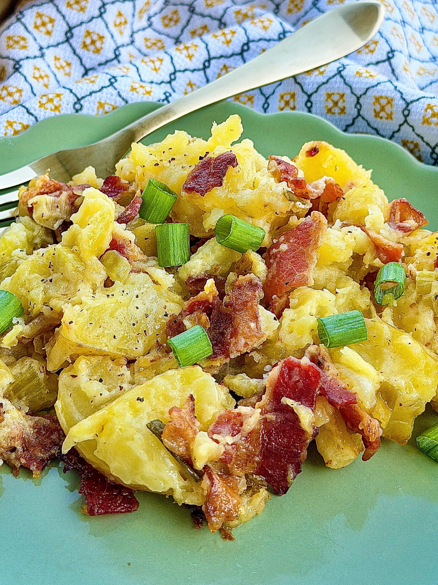 Grilled potato salad with cooked potatoes, crumbled bacon, chopped green onions, and pepper sitting on green scalloped plate with fork and towel in background
