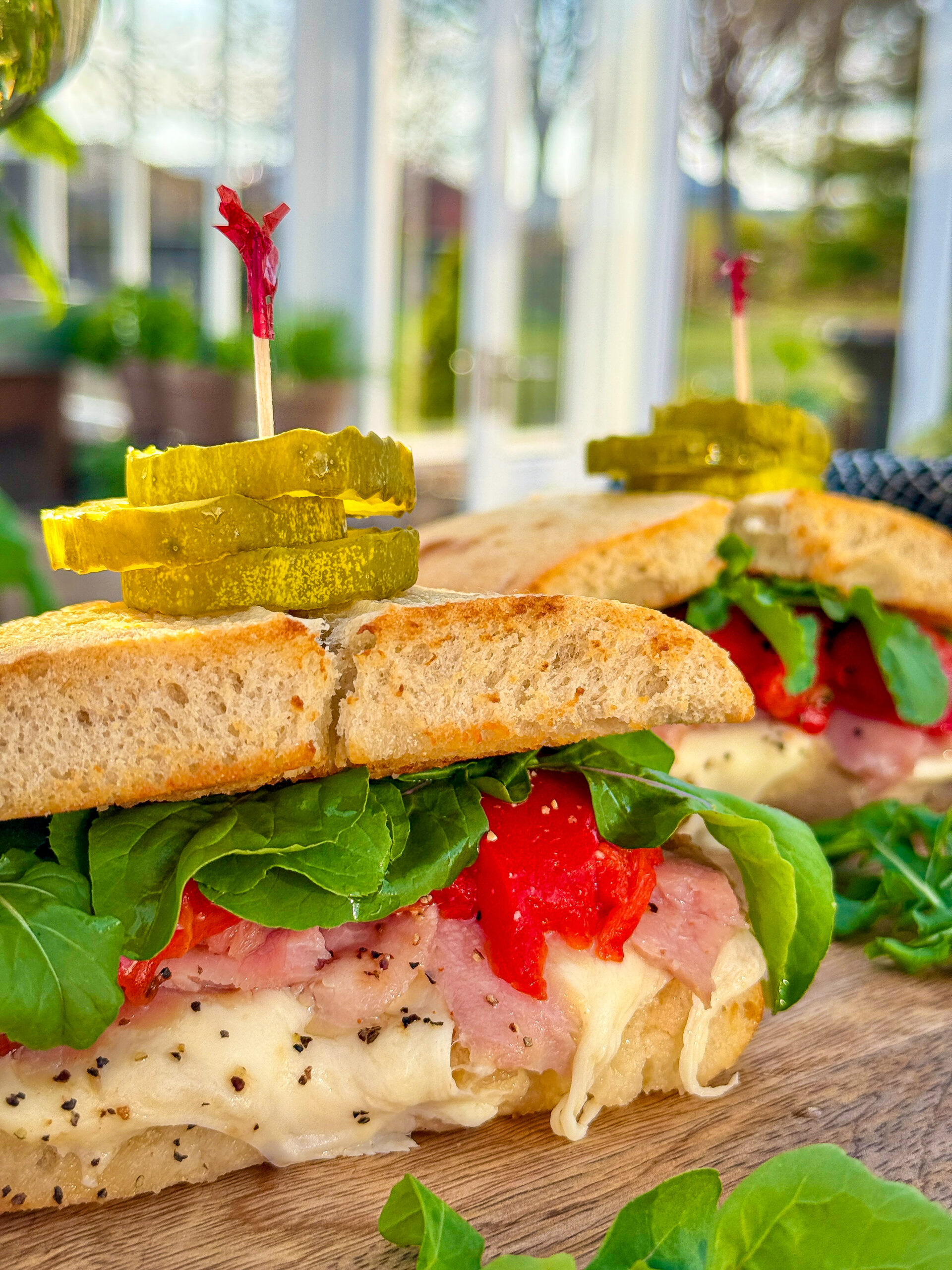 Ciabatta sandwiches with leafy greens, mozzarella, roasted red peppers, and ham, with pickle slices on top being held in with toothpicks sitting on brown table with white frame greenhouse blurred in background