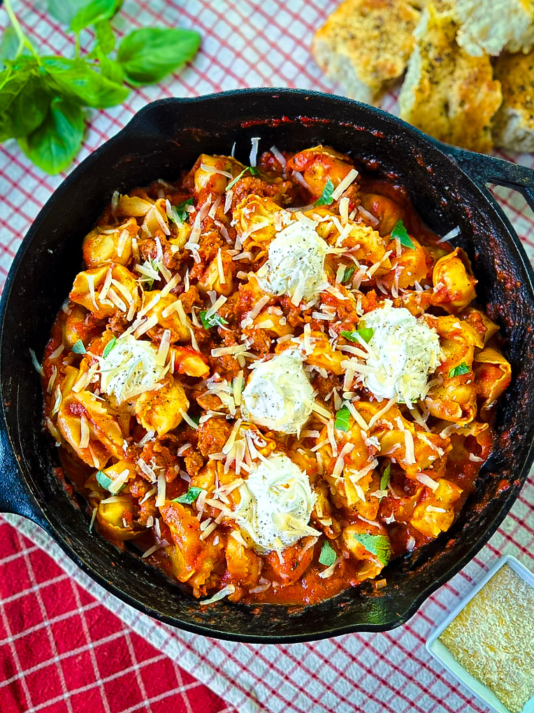 Black cast iron skillet with red sauce tortellini, Italian sausage, five dollops of mascarpone, shredded Parmesan cheese, chopped fresh basil leaves, sitting on white and red checkered linens