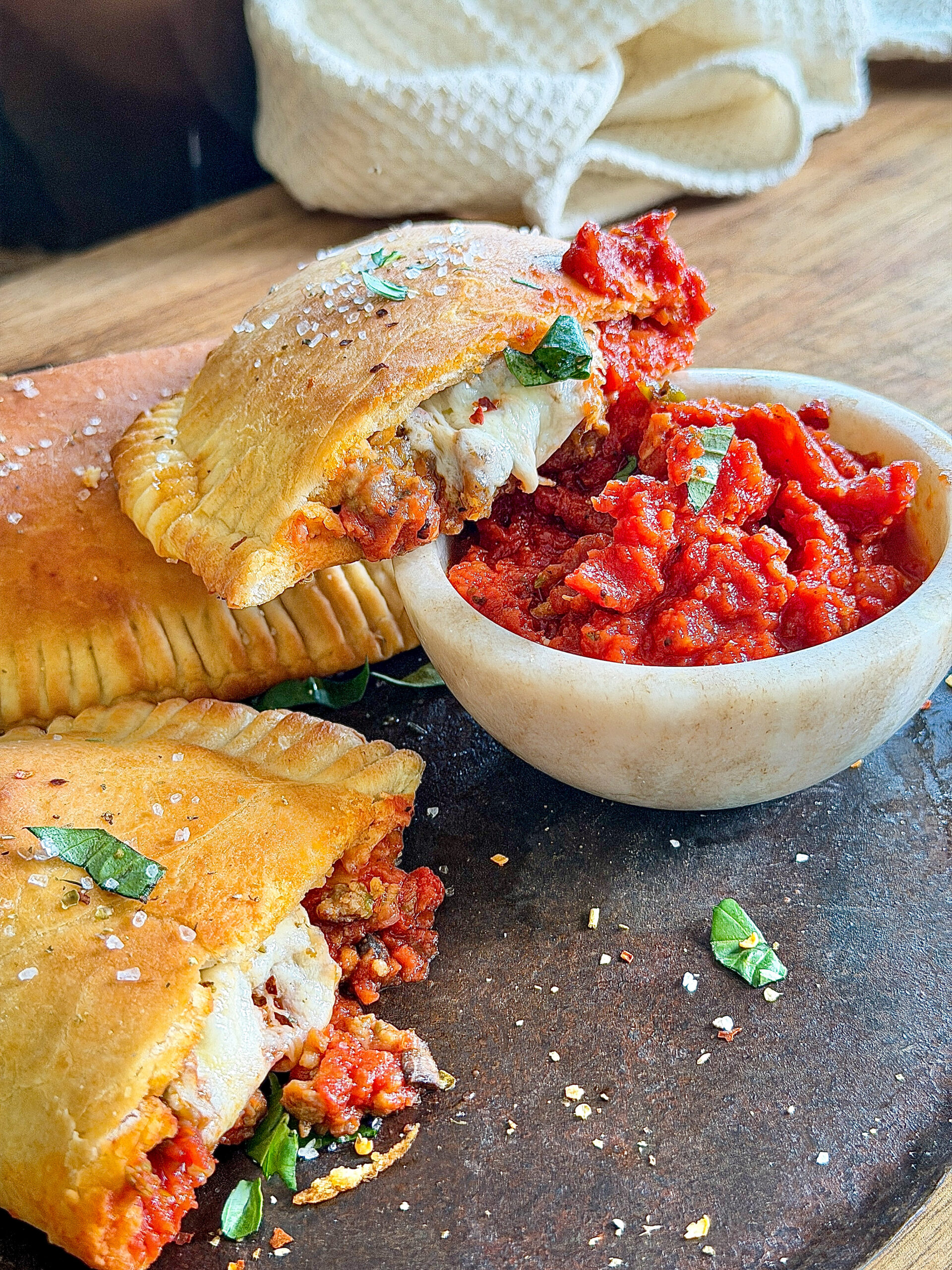 cream bowl of marinara with calzones sitting alongside, on top of black dish sitting on wood table with cream towel in background