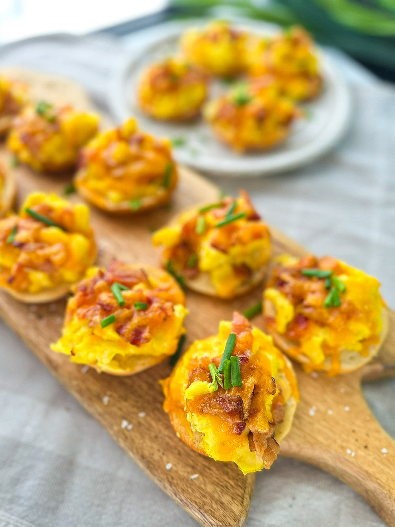 breakfast bagels with egg cheese bacon and chives sitting on brown cutting board sitting on cream towel with white plate of bagels in background