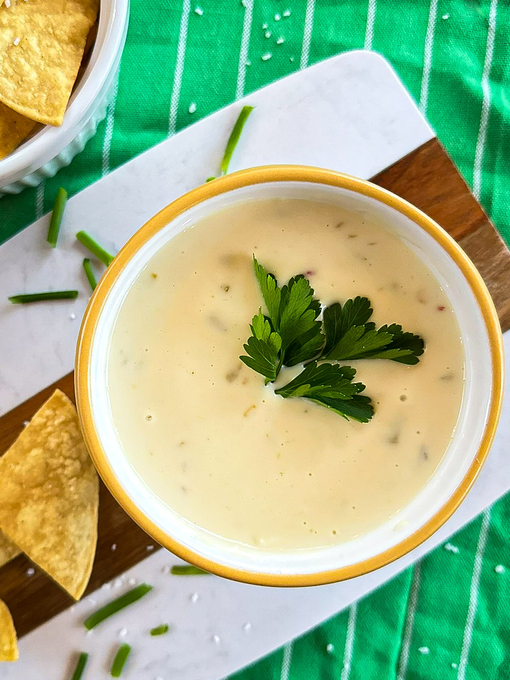 Bowl of queso cheese with green herbs sitting on brown and white cutting board with tortilla chips and herbs