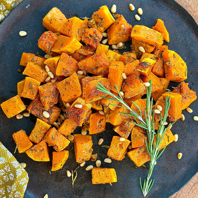 roasted butternut squash sprinkled with herbs and pine nuts sitting on a black plate with a sprig of fresh rosemary sitting on a brown wood table with a yellowish brown patterned towel