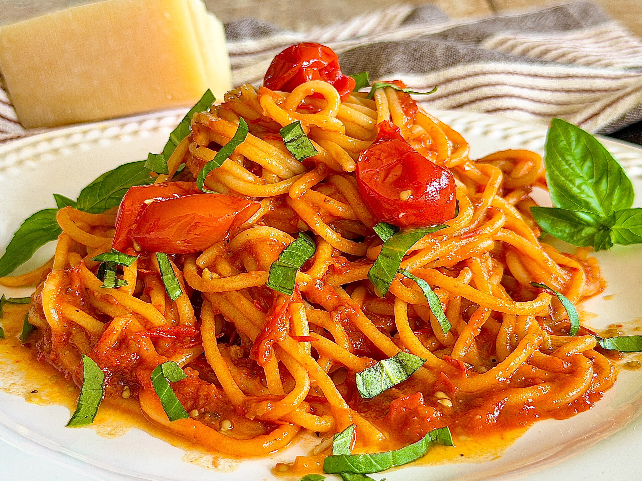 spaghetti noodles with roasted red cherry tomatoes and cut basil on a white plate with a block of parmesan cheese and a towel in the background
