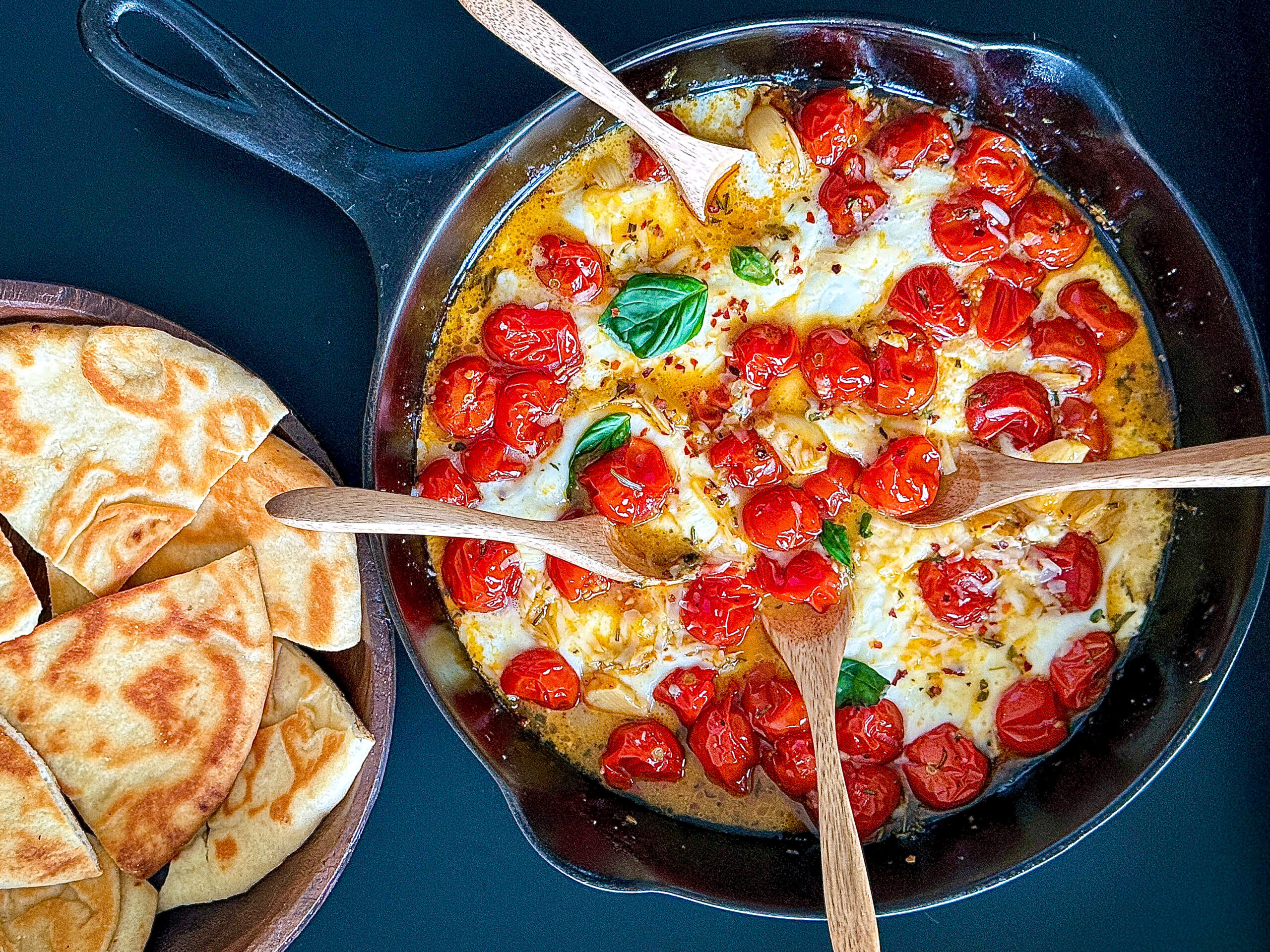 Cherry tomatoes in melted cheese in black cast iron skillet with wooden spoons and basil and toasted pita next to it on black surface