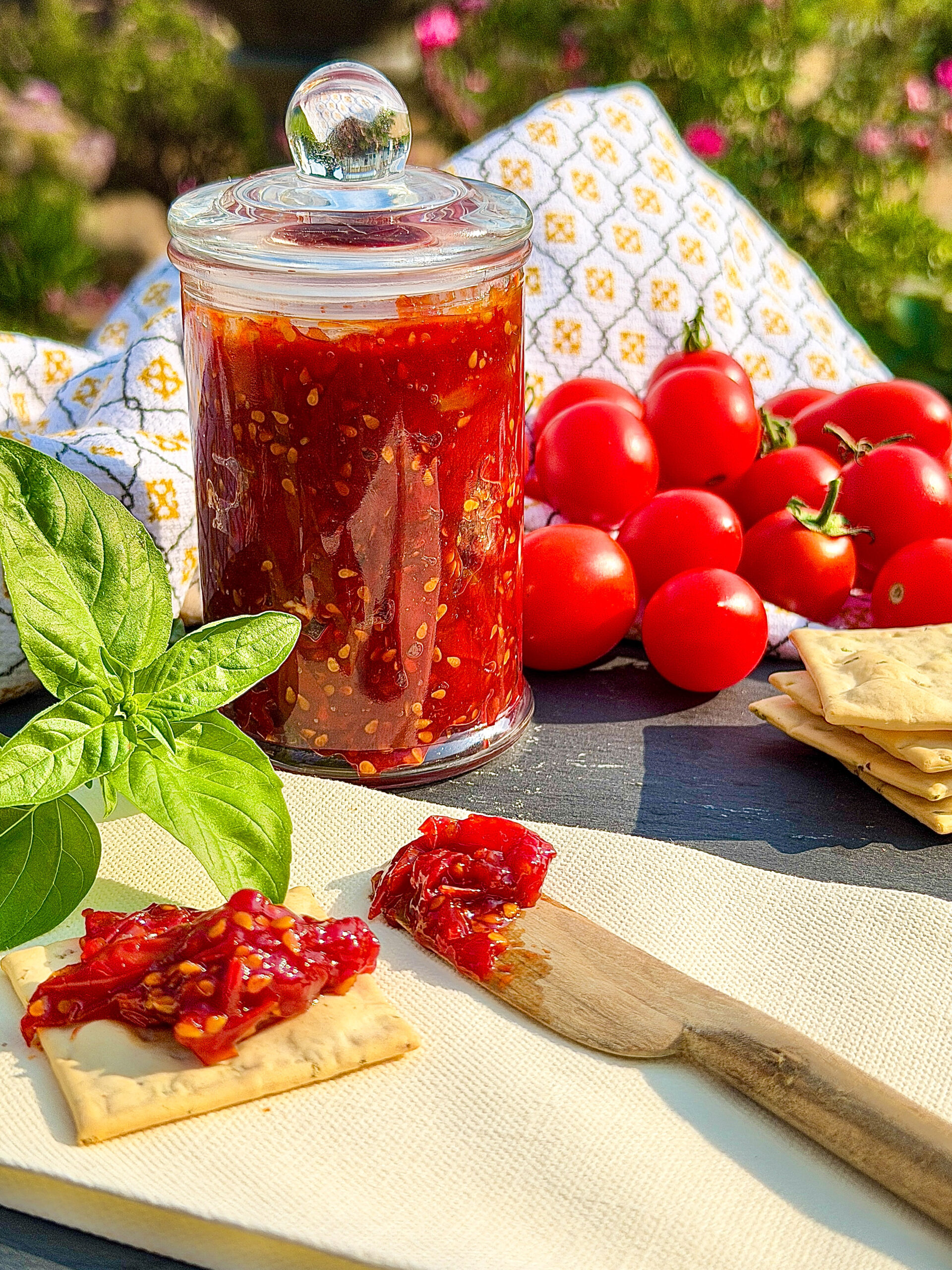 Glass jar of tomato jam sitting on gray slate table with a brown cutting board, wood knife, crackers, green basil, and patterned linen in background