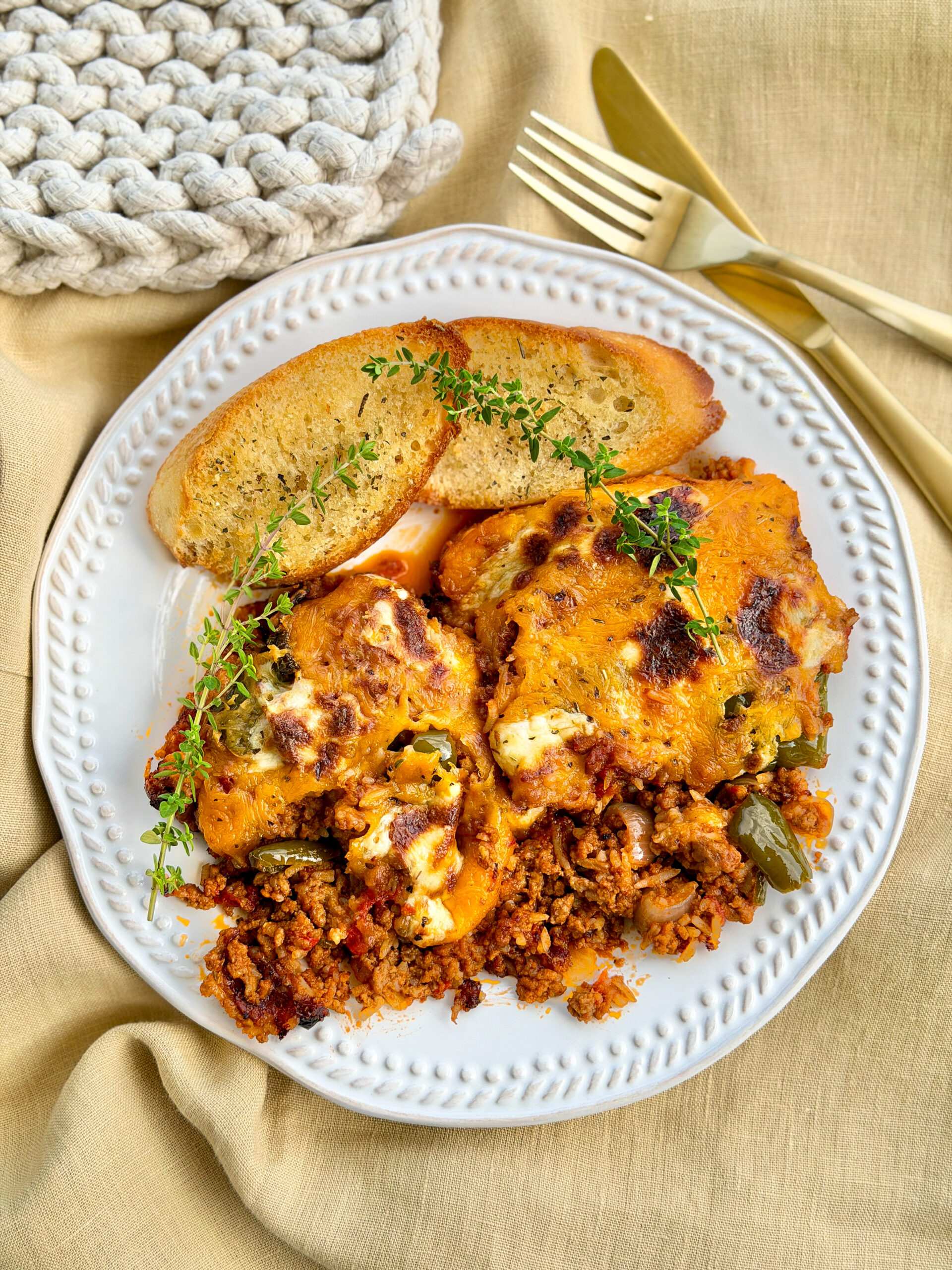 cheesy rice casserole with garlic bread and fresh herb sprigs sitting on white plate with gold fork and knife on top of tan linen and white crocheted pot holder