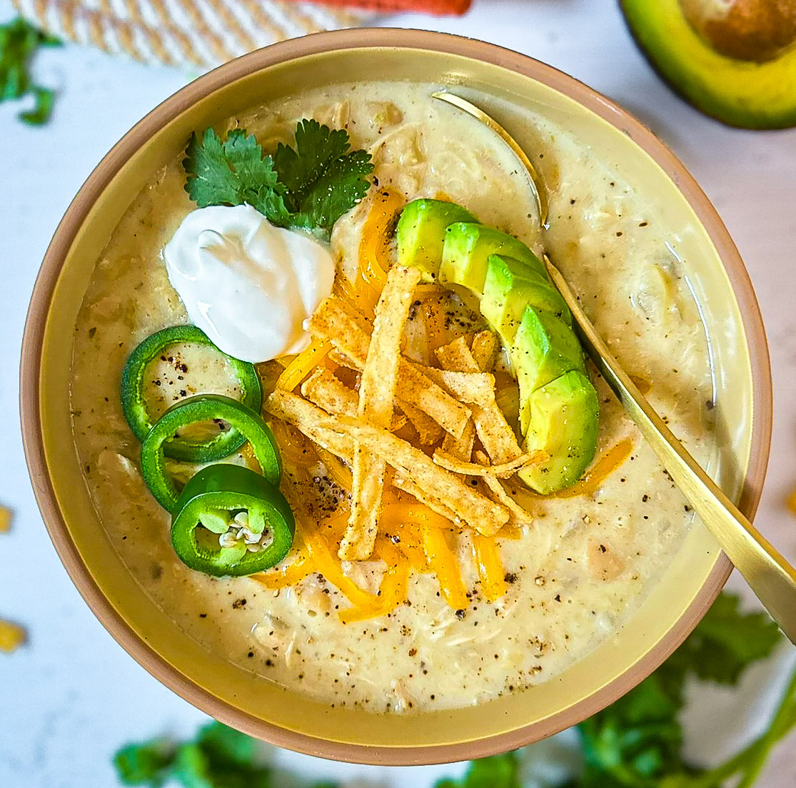 bowl of creamy soup with diced avocados, tortilla strips, shredded cheese, sliced jalapeños, sour cream, and cilantro, with gold spoon and with avocado cut in half and a green plant in background