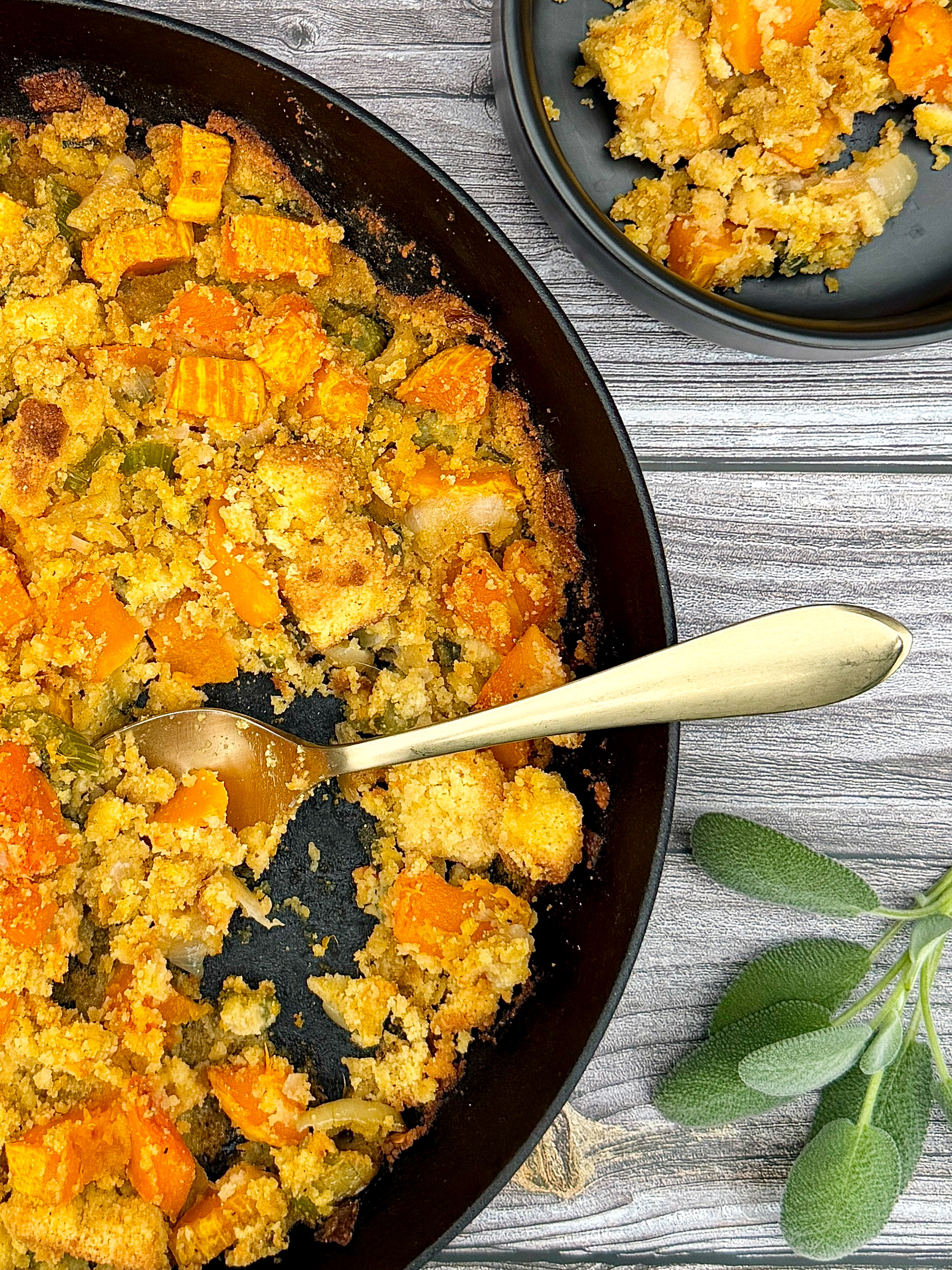 Butternut Squash Cornbread Casserole in black cast iron skillet with gold spoon, cream towel, sitting on gray wood surface with black plate of casserole next to it and greenery