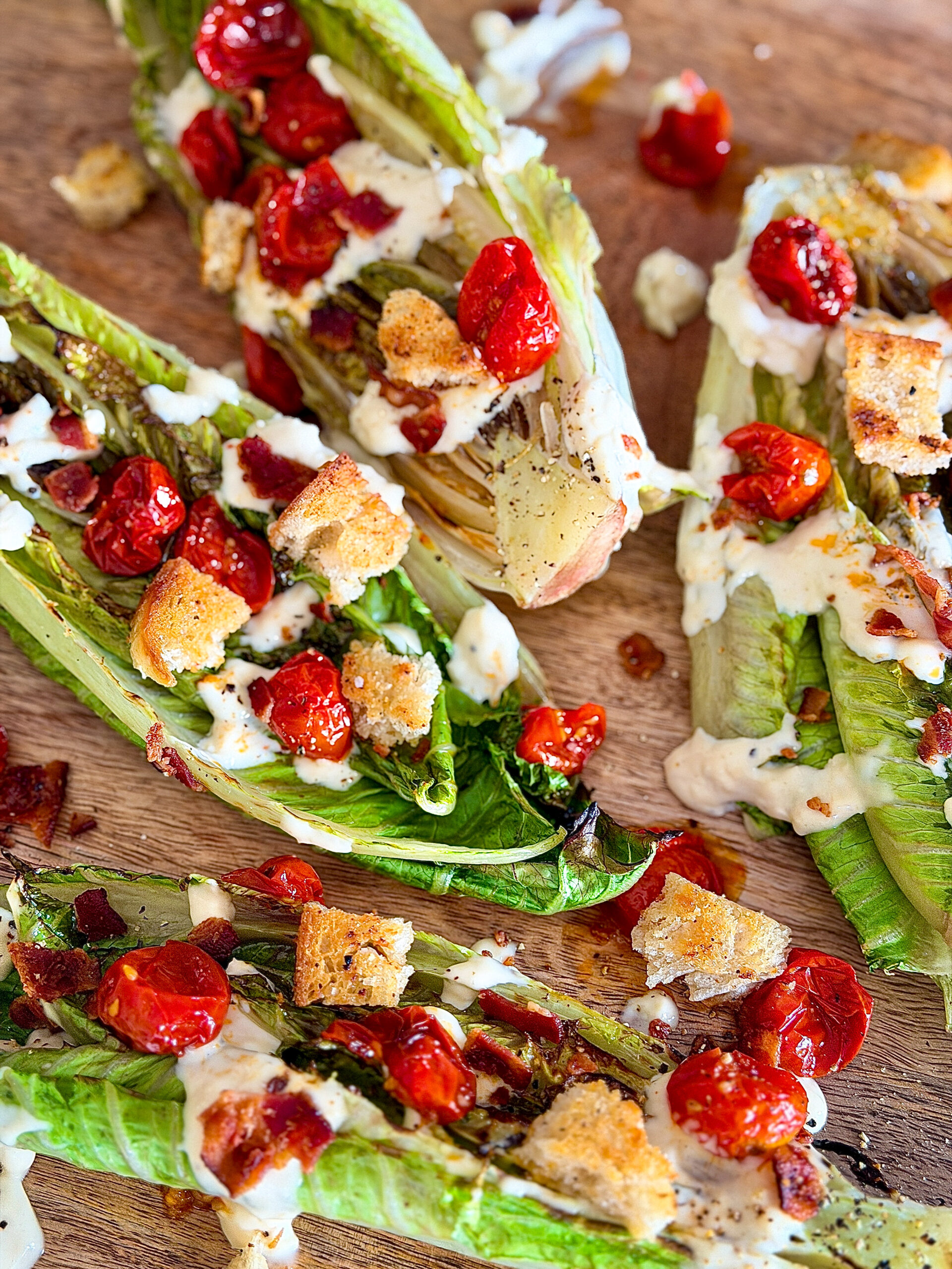 grilled romain lettuce halves with cooked cherry tomatoes, Caesar dressing drizzled, croutons, bacon, and seasonings on top of light wood surface