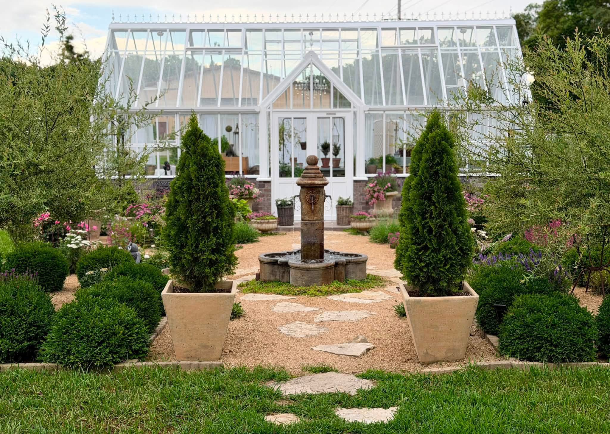 Hartley Greenhouse with fountain, stepping stones, potted evergreens, perennials, trees and potted urns at the door area