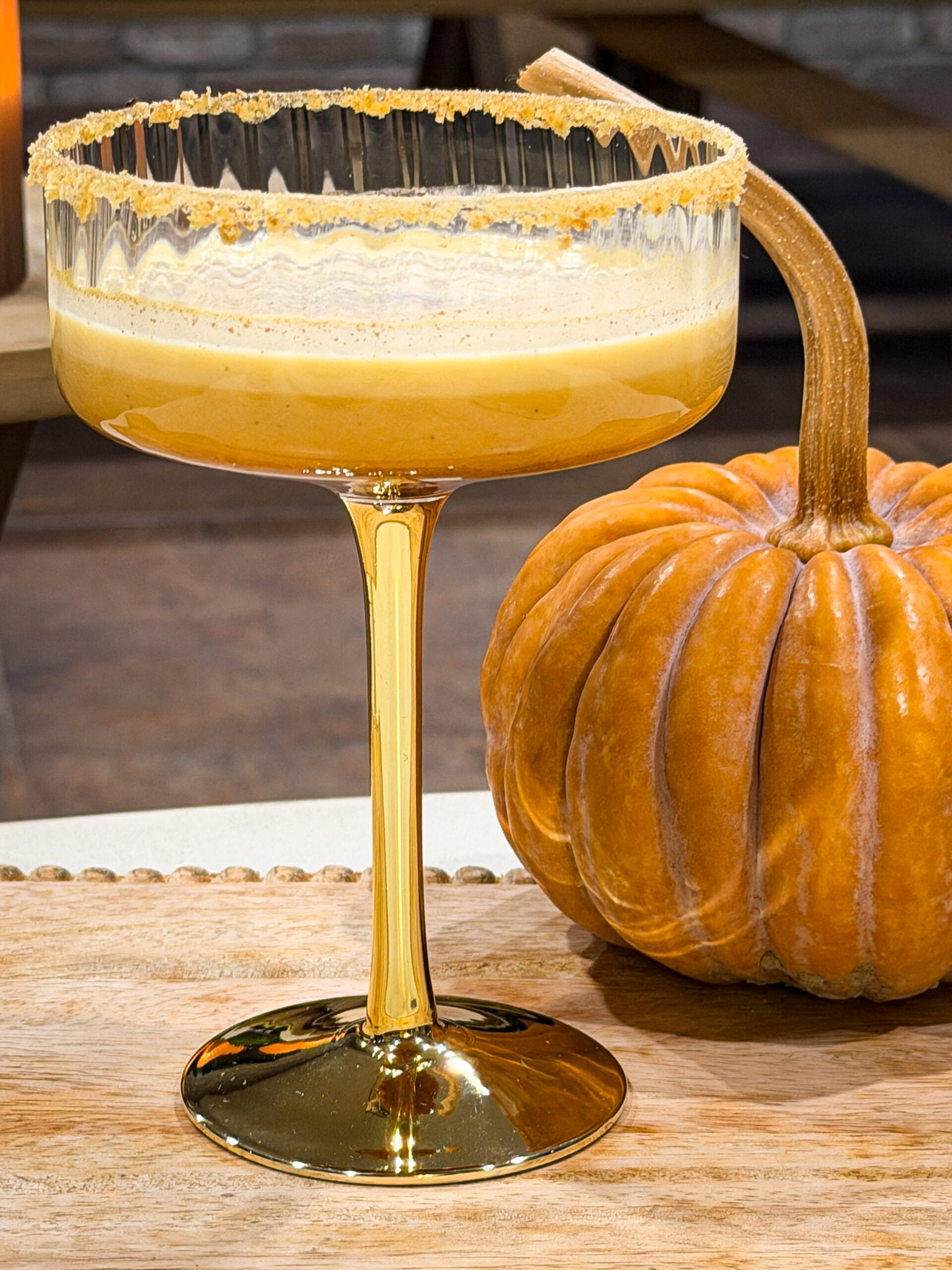 Pumpkin Spice Martini drink in gold stemmed martini glass with graham cracker crust rim, next to orange pumpkin decor sitting on brown wood surface