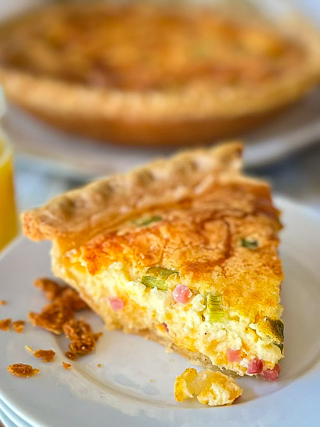 Slice of ham, egg, and green onion quiche sitting on white plates with juice and whole quiche in background